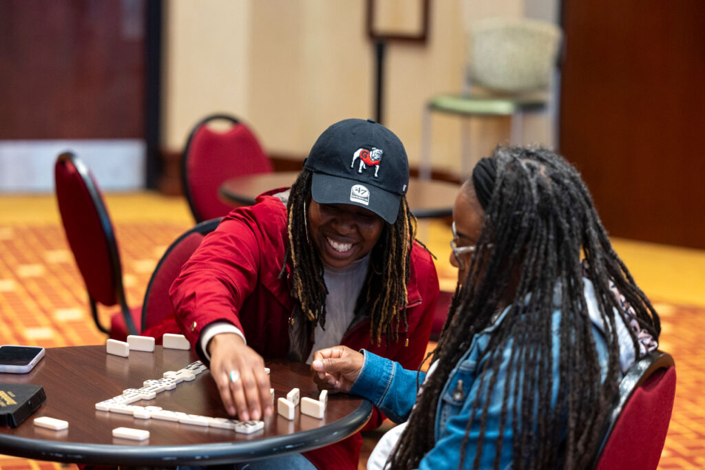 Two women smiling and playing dominoes at a round table in a carpeted room with red chairs.