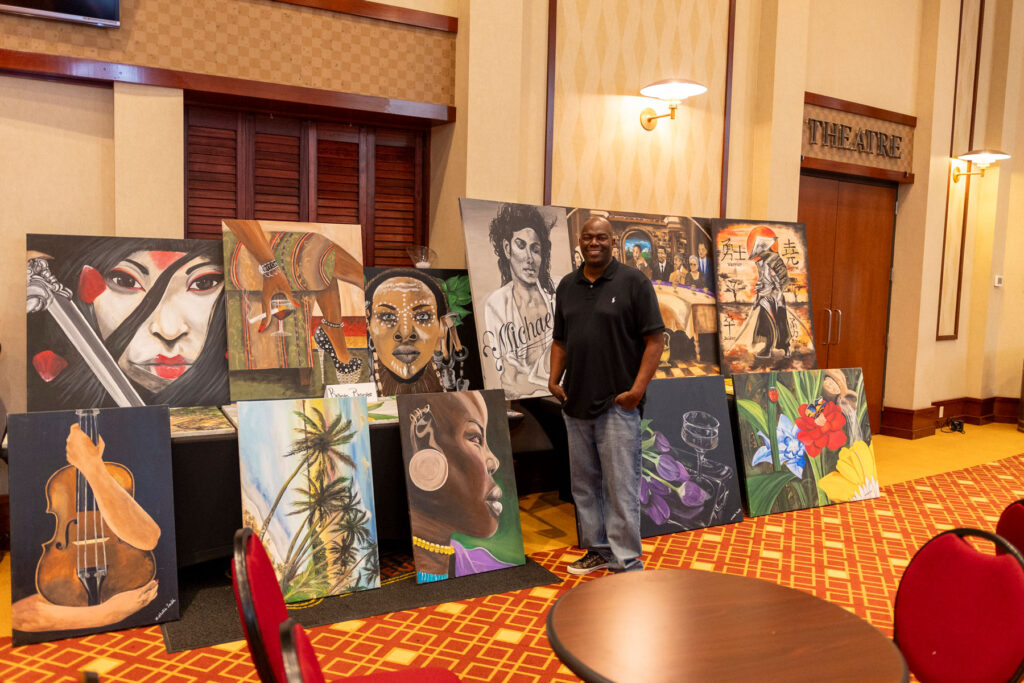 A man standing and smiling beside a display of colorful paintings featuring portraits, nature scenes, and cultural themes.