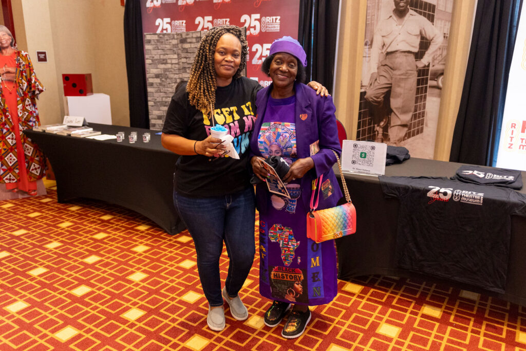 Two women posing together, one wearing a black graphic T-shirt and the other in a purple outfit decorated with Black history and African heritage designs.